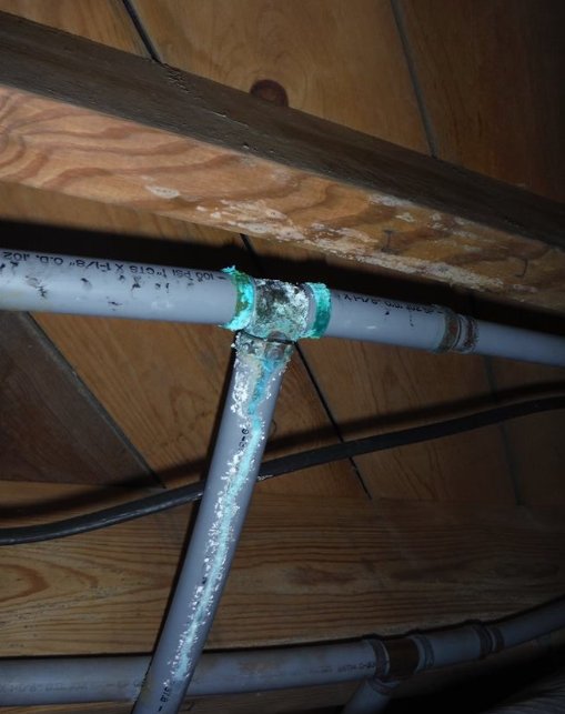 Gray polybutylene pipe with a corroded tee fitting showing green and white crusty buildup and mineral streaks
